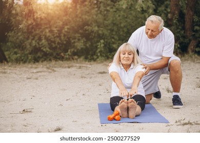 Senior couple engaging in yoga exercise outdoors, showcasing fitness, health, and togetherness in a natural setting. A man helps a woman do exercises. - Powered by Shutterstock