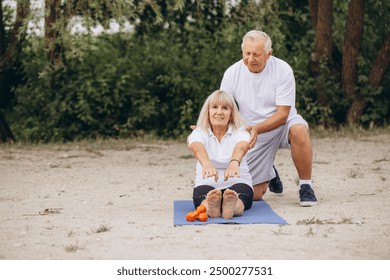 Senior couple engaging in yoga exercise outdoors, showcasing fitness, health, and togetherness in a natural setting. A man helps a woman do exercises. - Powered by Shutterstock