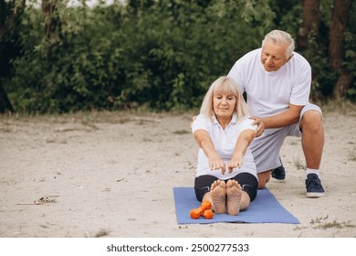 Senior couple engaging in yoga exercise outdoors, showcasing fitness, health, and togetherness in a natural setting. A man helps a woman do exercises. - Powered by Shutterstock