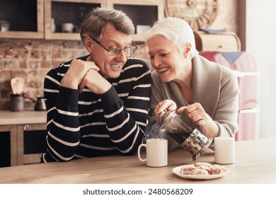 A senior couple is enjoying a cup of coffee and some snacks in a kitchen. The woman is pouring coffee from a French press into a mug for her husband, who is sitting across from her. - Powered by Shutterstock