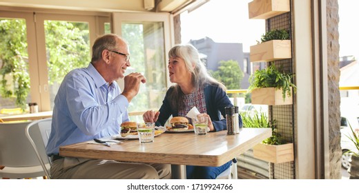 Senior Couple Enjoying Eating Burger Together At A Cafe. Smiling Mature Man And Woman Eating Burger At A Restaurant.