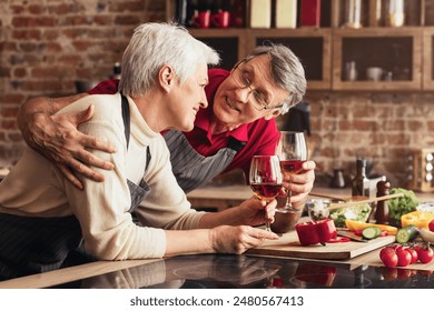 A senior couple is enjoying a glass of wine together in their kitchen. The man is leaning in towards his wife and smiling as they both raise their glasses. - Powered by Shutterstock