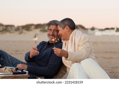 Senior couple enjoying picnic at seashore, holding sparklers and laughing. Lady with short hair and happy grey-haired man having fun together at sunset on the beach. Love, relationship concept - Powered by Shutterstock