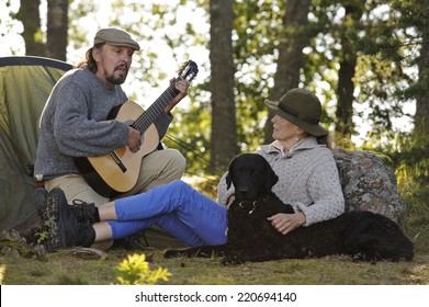 Senior couple enjoying some music outside their tent in evening light. Man plays guitar and they sing together. They have their pet curly coated retriever with them - Powered by Shutterstock