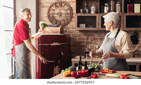 A senior couple enjoys preparing a meal together in a charming kitchen. Man, wearing a red shirt and an apron, stirs a bowl of salad, while the woman, also wearing an apron, opens the red refrigerator - Powered by Shutterstock