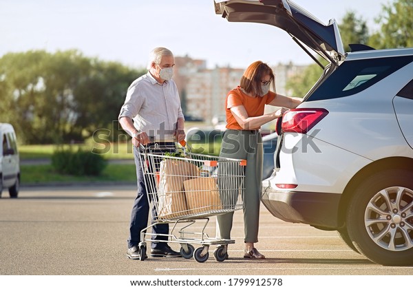 Senior couple\
with face masks putting shopping in car outside supermarket in city\
                                  \
