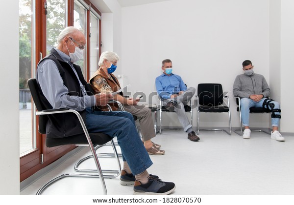 Senior couple with face masks sitting in a\
waiting room of a hospital together with a young and mature man -\
focus on the old man in the\
foreground