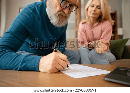 Senior Couple Filling In Form Signing Papers Sitting On Sofa Near Table Indoors. Paperwork Concept. Elderly Man Writing His Will In Notary Office. Cropped Shot, Selective Focus