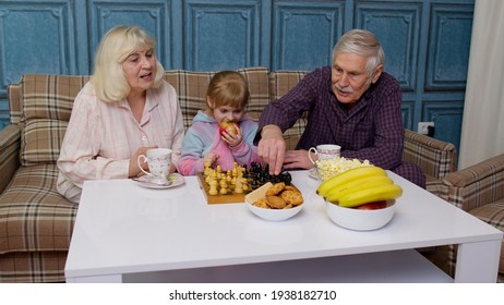 Senior Couple Grandparents With Child Granddaughter Spending Time Home Together, Sitting, Playing Chess Game With On Table In Living Room. Old Grandmother, Grandfather With Cute Girl Grandkid Girl Kid