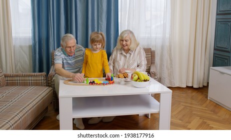 Senior Couple Grandparents With Child Kid Granddaughter Spending Time Home Together, Sitting, Playing Game, Riding Toy Train On Wooden Railway On Table. Old Grandmother Grandfather With Girl Grandkid