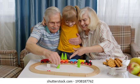 Senior Couple Grandparents With Child Kid Granddaughter Spending Time Home Together, Sitting, Playing Game, Riding Toy Train On Wooden Railway On Table. Old Grandmother Grandfather With Girl Grandkid