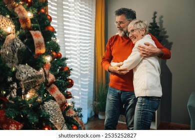 Senior couple happily celebrating christmas at home, embracing in front of a decorated tree, surrounded by festive lights and ornaments, creating a cozy atmosphere - Powered by Shutterstock