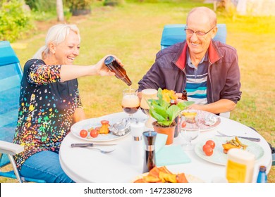 Senior Couple Having Barbecue Dinner Outside - Happy Woman Pouring Beer In The Glass Sitting At The Table - Two Smiling Retirees Having Fun Eating Outdoors