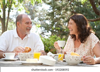 Senior Couple Having Breakfast Together At A Table In A Luxury Hotel Garden During A Sunny Day. Mature Joyful People Eating Healthy Food And Having Fun While Drinking Coffee. Outdoors Lifestyle.