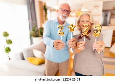 Senior couple having fun spending winter holiday season together at home, holding small golden balloons shaped as numbers 2023, representing upcoming New Year - Powered by Shutterstock