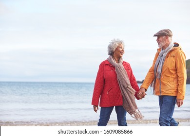 Senior Couple Hold Hands As They Walk Along Shoreline On Winter Beach Vacation