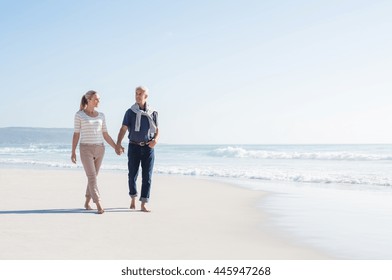 Senior couple holding hands at the beach on a sunny day. Mature couple in love holding hands and looking each other at the seaside. Smiling wife and happy husband walking barefoot on the white sand.