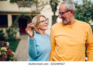 Senior couple holding up new house key while standing outside. Portrait of elderly mature caucasian husband and wife feeling happy about buying a new house. Focus on a keys in woman hands. - Powered by Shutterstock