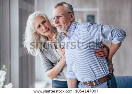Senior couple at home. Handsome old man is having back pain and his attractive old woman supports him.