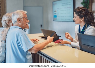 A senior couple at the hotel front desk makes contactless payments with a smartphone. Seniors traveling and using new technologies - Powered by Shutterstock