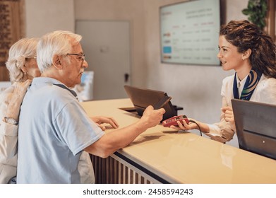 A senior couple at the hotel front desk makes contactless payments with a smartphone. Seniors traveling and using new technologies - Powered by Shutterstock