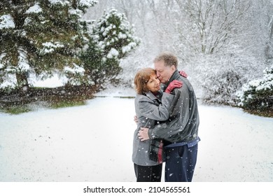 Senior Couple Hugging Outdoors In Snowfall On Valentine's Day In Midwest; Man Kissing Woman