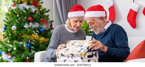 Senior couple husband and wife opening gift present box, sitting on sofa in room with Christmas tree and decoration - Powered by Shutterstock