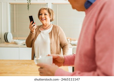 Senior couple in a kitchen with a mug on the table beside them. Smiling senior woman looking at her phone, and senior man using a digital tablet.  - Powered by Shutterstock