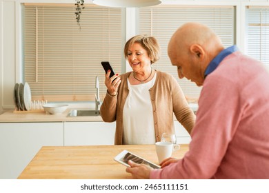 Senior couple in a kitchen with a mug on the table beside them. Smiling senior woman looking at her phone, and senior man using a digital tablet.  - Powered by Shutterstock