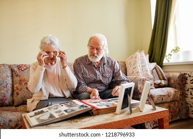 Senior Couple Looking At Family Photo Album With Vintage Photos Sitting On The Sofa In Living Room With Soft Cozy Interior, Old Lady Points At Photos And Tells Different Stories To Her Husband.