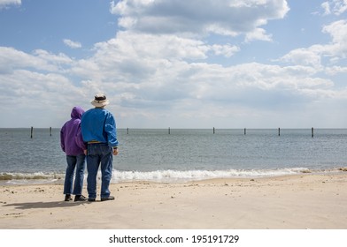 A Senior Couple Looking Out At The Ocean On A Sandy New England Beach.