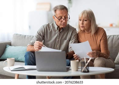 Senior Couple Looking Through Papers Using Laptop Reading Bills And Counting Expenses Together Sitting On Couch At Home. Household And Finances, Retirement Lifestyle Concept