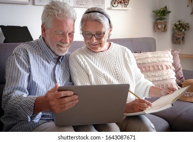 A Senior Couple Looks At The Laptop With Interest And Makes A Note In A Notebook. Plan A Trip. White Hair. New Technology For Older People