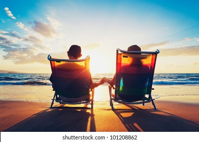 Senior Couple Of Old Man And Woman Sitting On The Beach Watching Sunset