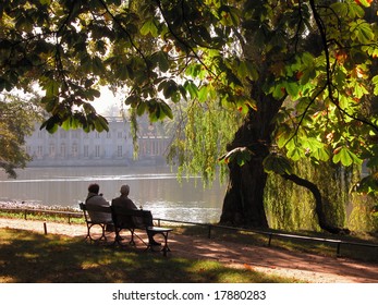 Senior Couple On A Late Summer Day, Enjoying The Silence. Seniors Sitting On A Park Bench And Looking At The Palace On The Water.