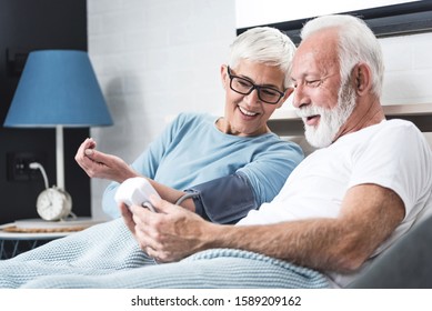 Senior Couple In Pajamas In A Bed In A Bedroom Measure Each Other's Blood Pressure. Concept Of Home Health Check