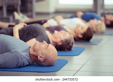 Senior couple participating in aerobics class at the gym with diverse people lying in a receding row on mats, focus to the couple in the foreground, healthy lifestyle concept