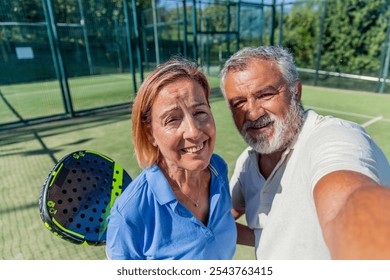 senior couple pauses mid-match to take a cheerful selfie together on the padel court, capturing the fun and excitement of their game - Powered by Shutterstock