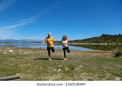 Senior couple practicing leg stretches by a tranquil lake, enjoying a healthy and active lifestyle amidst nature's beauty on a sunny day - Powered by Shutterstock