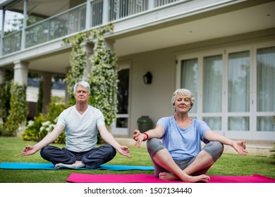 Senior Couple Practising Yoga On Exercise Mat In Lawn. They Are Meditating In The Lotus Position