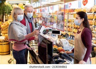 Senior Couple In Protective Face Masks Paying At Cashdesk At Supermarket, Elderly Family Shopping While Coronavirus Pandemic, Cashier Standing Behind Checkout Screen. Shopping While Quarantine