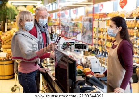Senior couple in protective face masks paying at cashdesk at supermarket, elderly family shopping while coronavirus pandemic, cashier standing behind checkout screen. Shopping while quarantine