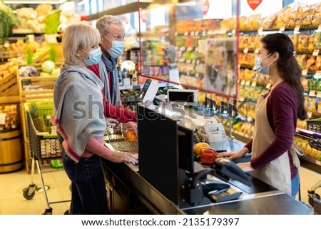 Senior couple in protective face masks paying at cashdesk at supermarket, elderly family shopping while COVID-19 pandemic, cashier standing behind checkout screen. Shopping while quarantine concept