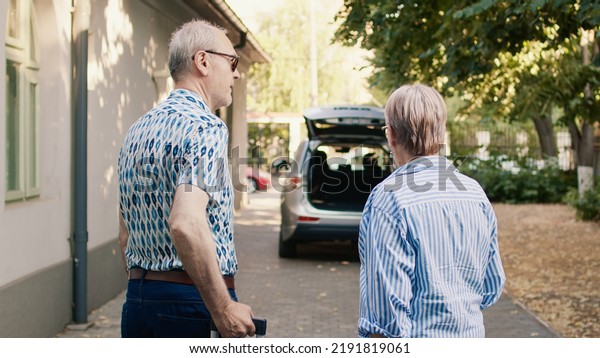 Senior couple putting luggage\
in car trunk while getting ready for holiday trip. Cheerful elders\
going on retirement voyage while having heavy baggage and travel\
trolley.