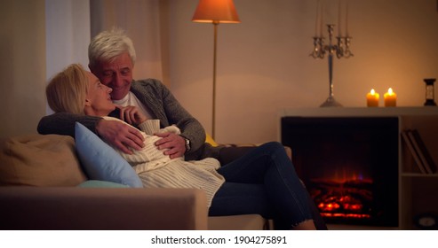 Senior Couple Relaxing And Talking Together Sitting On Sofa In Living Room At Home. Side View Of Happy Aged Man And Woman Sitting On Couch Near Fireplace And Chatting Enjoying Evening At Home