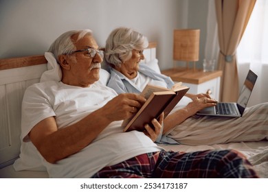 Senior couple relaxing in their bed. Woman is using laptop while man is reading book.  - Powered by Shutterstock