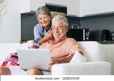 Senior Couple Relaxing Together In The Kitchen With A Laptop And With A Cup Of Coffee Or Tea. She Caresses Him And Smiling Happily Pass Together. Everyday Real Life At Home For Mature Retired People