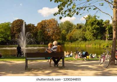 Senior Couple Resting On The Bench At Park With Some Young People And Bikes As An Autumn Scene.