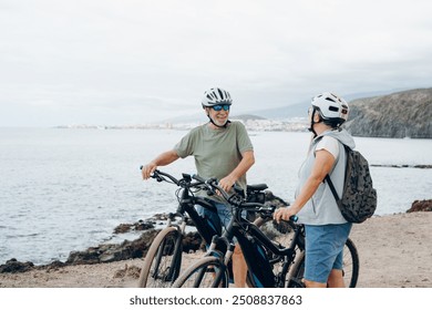 Senior couple riding bikes together in rocky beach enjoying outdoor. Active mature people talking holding mountain bikes. Bicycle lifestyle concept, having fun - Powered by Shutterstock