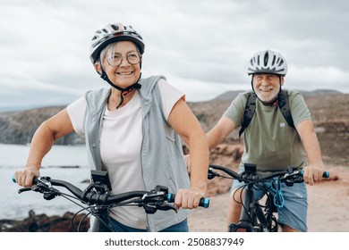 Senior couple riding bikes together in rocky beach enjoying outdoor. Active mature people talking holding mountain bikes. Bicycle lifestyle concept, having fun - Powered by Shutterstock